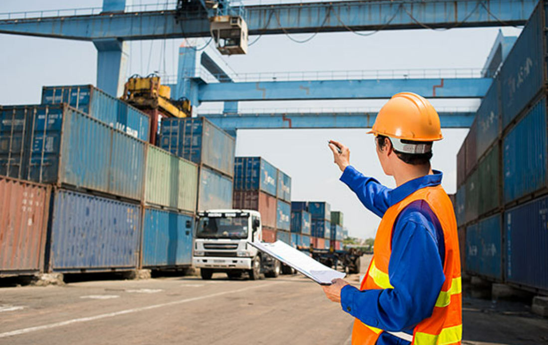 A close-up of a cargo ship loaded with multiple colorful shipping containers, docked at a busy port. In the background, cranes are actively unloading cargo, highlighting the bustling nature of international trade. The clear blue sky and calm water reflect a sense of security and reliability. This image represents Monoline Cargo Insurance provided by the Insurance Company Worthington Ohio LLC, ensuring safe and protected transportation of goods.